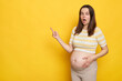 Shocked surprised Caucasian pregnant woman in casual clothing posing isolated over yellow background pointing away at copy space for advertisement or promotional text
