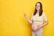 Happy joyful Caucasian pregnant woman in casual clothing indicating at area for advertising content empty space for promotion posing isolated over yellow background