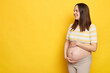 Cheerful delighted brown haired pregnant woman with bare belly posing isolated over yellow background stroking her belly looking away at copy space for advertisement