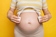 Unrecognizable young Caucasian pregnant woman dressed in top posing against bright yellow wall holding headphones future baby listening to classic music for development