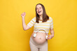Overjoyed excited Caucasian pregnant woman in casual clothing posing isolated over yellow background dancing with raised arm holding headphones near her big bare belly