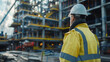 © Nano Photos - A construction worker in a yellow high-visibility jacket and a white safety helmet observes a large construction site with cranes and scaffolding.