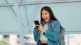 Fototapeta  - Young asian woman using mobile in city. Happy female tourist wearing jeans jacket and holding smartphone at public