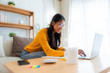 Fototapeta  - Young Asian female wearing glasses using laptop, working at home in living room, coffee mug on table. Cozy office workplace, remote work
