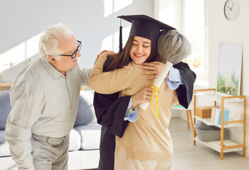 Portrait of a happy smiling graduate girl student hugging with her senior old parents holding diplomas in hands and celebrating university graduation wearing graduate gown at home.