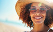 A woman with curly hair and sunglasses is smiling and wearing a straw hat
