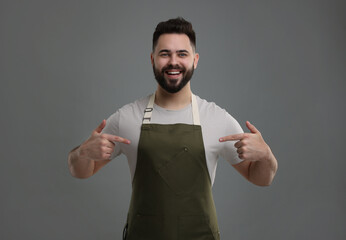 Canvas Print - Smiling man pointing at kitchen apron on grey background. Mockup for design