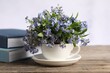 Beautiful forget-me-not flowers in cup, saucer and books on wooden table against light background, closeup