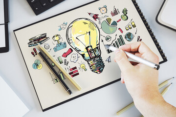 A person's hand drawing a creative concept with a glowing light bulb on paper, symbols and graphs, on a white desk