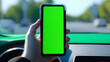 smartphone with blank green screen held by hand inside a moving car, presenting setting perfect for navigation or travel app promotions. The blurred dashboard and road in the background