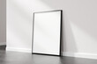 Blank picture frame with a black border leaning against a white wall in a room with light hardwood flooring, 3d render.