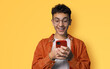 Smiling funny young man wear braces, sunglasses glasses, orange denim shirt hold typing text look read cell phone cellular smartphone cellphone, isolated against yellow background.