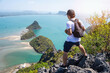 A male hiker with a backpack stands on a rocky peak, enjoying the expansive view of a serene tropical bay and islands.