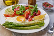 Round plate with cooked asparagus, fried egg, avocado and fresh vegetable salad on the table