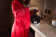 An Asian woman in her red satin nightgown is standing, pouring hot water into a white cup in the middle of the night.