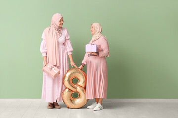 Poster - Muslim adult woman with her mother, gift boxes and balloon in shape of figure 8 near green wall. International Women's Day celebration