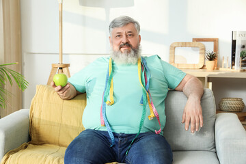 Canvas Print - Overweight happy mature man with measuring tapes and apple sitting on sofa at home. Weight loss concept