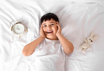 Sticker - Cute happy little boy with toy rabbit and alarm clock lying in bedroom