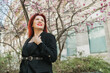 Fashion outdoor photo of beautiful woman with red curly hair in elegant suit posing in spring flowering park with magnolias tree. Copy space and empty place for advertising text