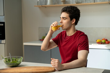 A man is seated at a table, holding a cup and drinking orange juice. He seems focused as he takes a sip from the glass.