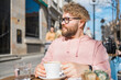 Young bearded man having breakfast at table of street cafe on spring day, drinking warm cappuccino and enjoy morning.