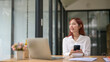 © apichat - A woman is sitting at a desk with a laptop and a cell phone. She is smiling and she is enjoying her time