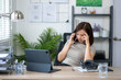 © Wasana - A woman is talking on her cell phone while sitting at a desk. She is wearing a gray shirt and has her head down. The desk is cluttered with papers and a laptop