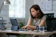 © Wasana - A woman is sitting at a desk with a laptop and a glass of water. She is focused on her work and she is in a serious mood