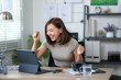 © Wasana - A woman is sitting at a desk with a laptop and a tablet. She is smiling and she is happy