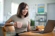 © Wasana - A woman is sitting at a table with a laptop and a pink mug. She is smiling and she is enjoying her time