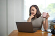 © Wasana - A woman is sitting at a table with a laptop and a pink mug. She is looking at the laptop and seems to be thinking