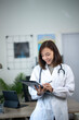 © Wasana - A woman in a white lab coat is smiling as she writes on a clipboard. She is in a medical setting, possibly a doctor's office or hospital