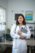 © Wasana - A woman in a white lab coat is holding a tablet and smiling. She is standing in front of a desk with a computer and a potted plant
