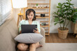 © Wasana - A woman is sitting on a couch with a tablet in her lap. She is wearing headphones and she is focused on her device. The room is decorated with a potted plant and a vase