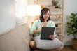 © Wasana - A woman is sitting on a couch with a tablet in her lap. She is wearing headphones and she is focused on the screen. The room is decorated with plants and has a cozy atmosphere