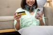 © Wasana - A woman is sitting on the floor with a credit card in her hand and a cell phone in her other hand. She is smiling and she is happy
