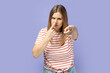 Suspicious blond woman in T-shirt pointing to camera, touching nose, doing liar gesture, body language symbol of cheats, falsehood and deception. Indoor studio shot isolated on purple background.