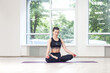 Full length portrait of calm realxed young woman doing yoga sitting in lotus pose meditating with closed eyes, wearing black pants and top. Indoor shot in gym near big windows.