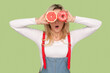 Portrait of shocked surprised adult blond woman standing covering eyes with grapefruit and doughnut, keeps mouth open, wearing denim overalls. Indoor studio shot isolated on light green background