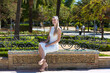 Beautiful young woman with long blonde hair and a short white dress is sitting on a bench in a famous park in Seville rich in vegetation and birds. Travel and holiday concept.
