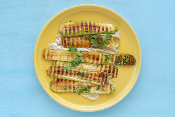Wall Mural - Grilled zucchini served with tahini pasta and chopped herbs on yellow platter over wooden background..