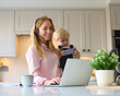 Busy Working Mother With Young Son At Home In Kitchen Using Laptop As Boy Plays With Her Phone