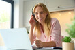 Woman Wearing Headset Working From Home In Kitchen Using Laptop
