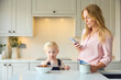 Mother At Home Looking At Mobile Phone As Young Son Eats Breakfast In Kitchen