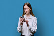 Teenage student girl in white shirt holding smartphone in hands on blue studio background