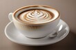 A cup of coffee sits on a saucer with a spoon beside it. The cup is white with a brown design on top. The coffee has a leaf pattern in the foam. The background is a brown color.