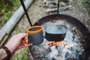 Sticker - Tourist mug for tea, man holding a glass in his hand on the background of a fire, tourist utensils for food, camping in nature.