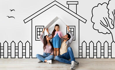 Happy middle eastern family of three father mother and daughter sitting on floor over illustrated house of their dreams over white wall background, parents holding a roof