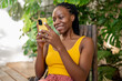 Cheerful african girl with smartphone. Smiling joyful black African American woman using phone resting among green plants leaves. Woman blogger writes blog posts, flips through feed on social networks