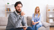 A bearded man therapist, holding a notebook, is seated attentively with a contemplative expression, while a lady client sits in the background on a couch